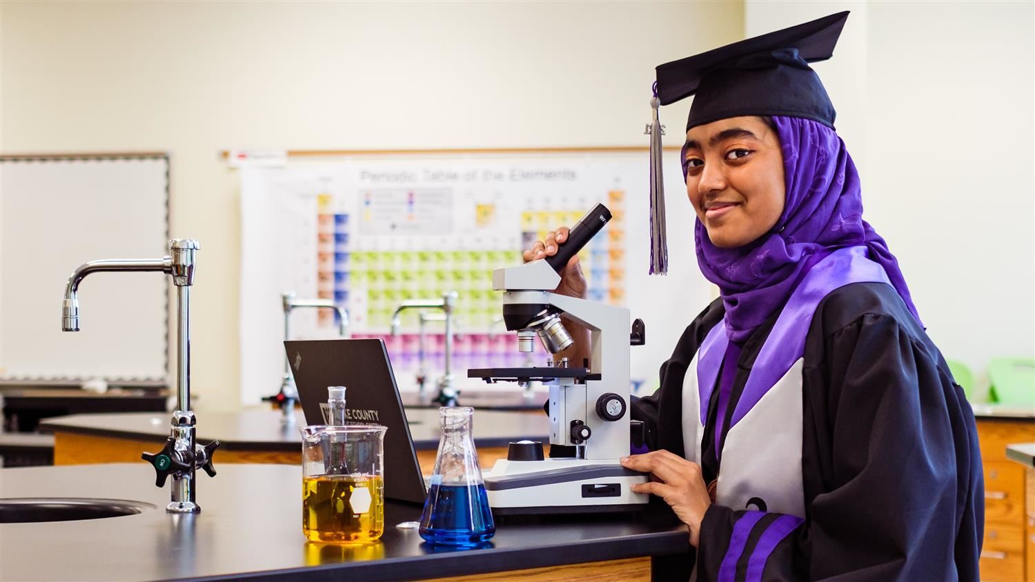 student wearing cap and gown using a microscope and working on a laptop while smiling