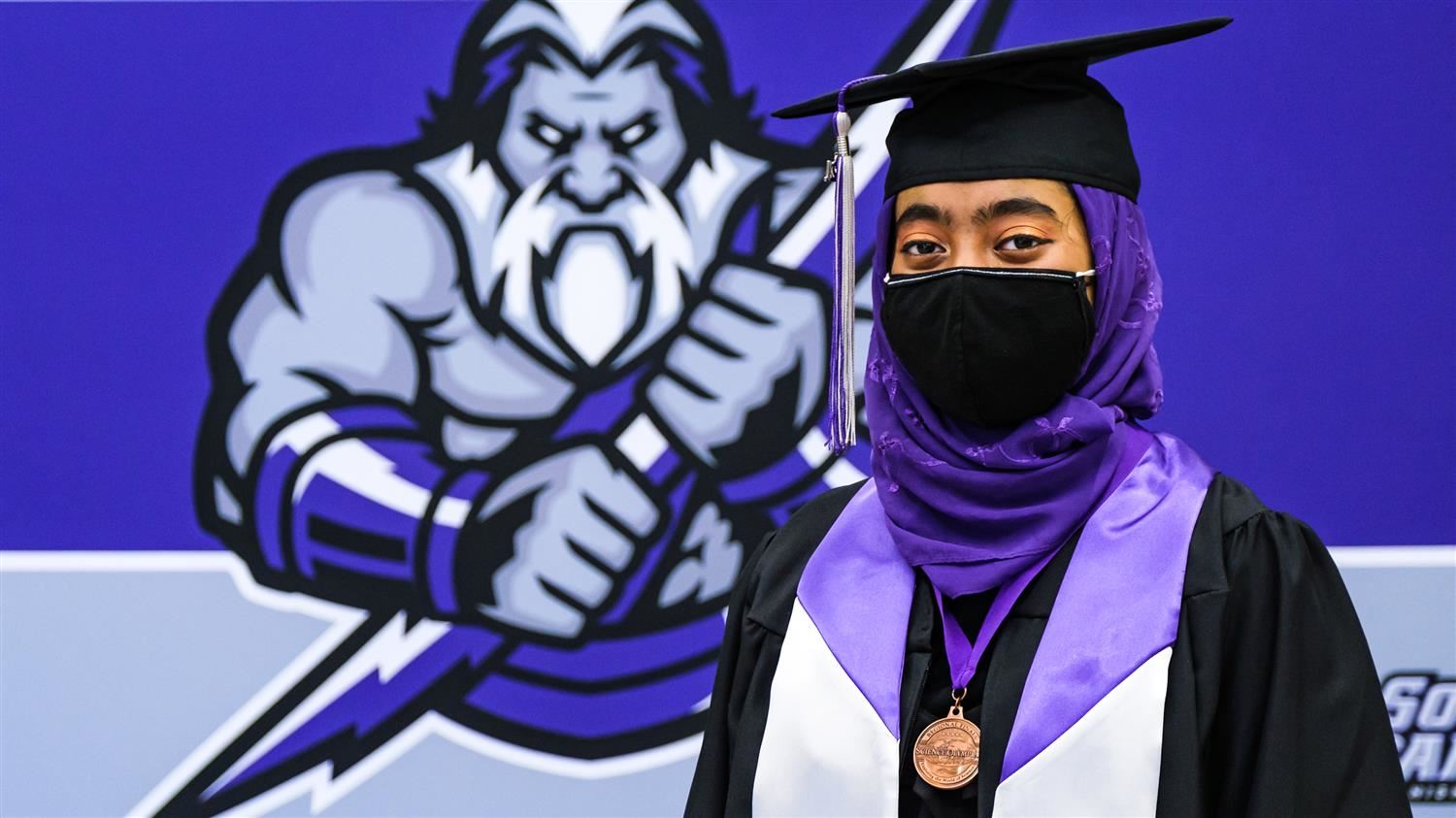 Student wearing a hijab, cap, gown, and medal with the South Garner High School Titan logo in the background