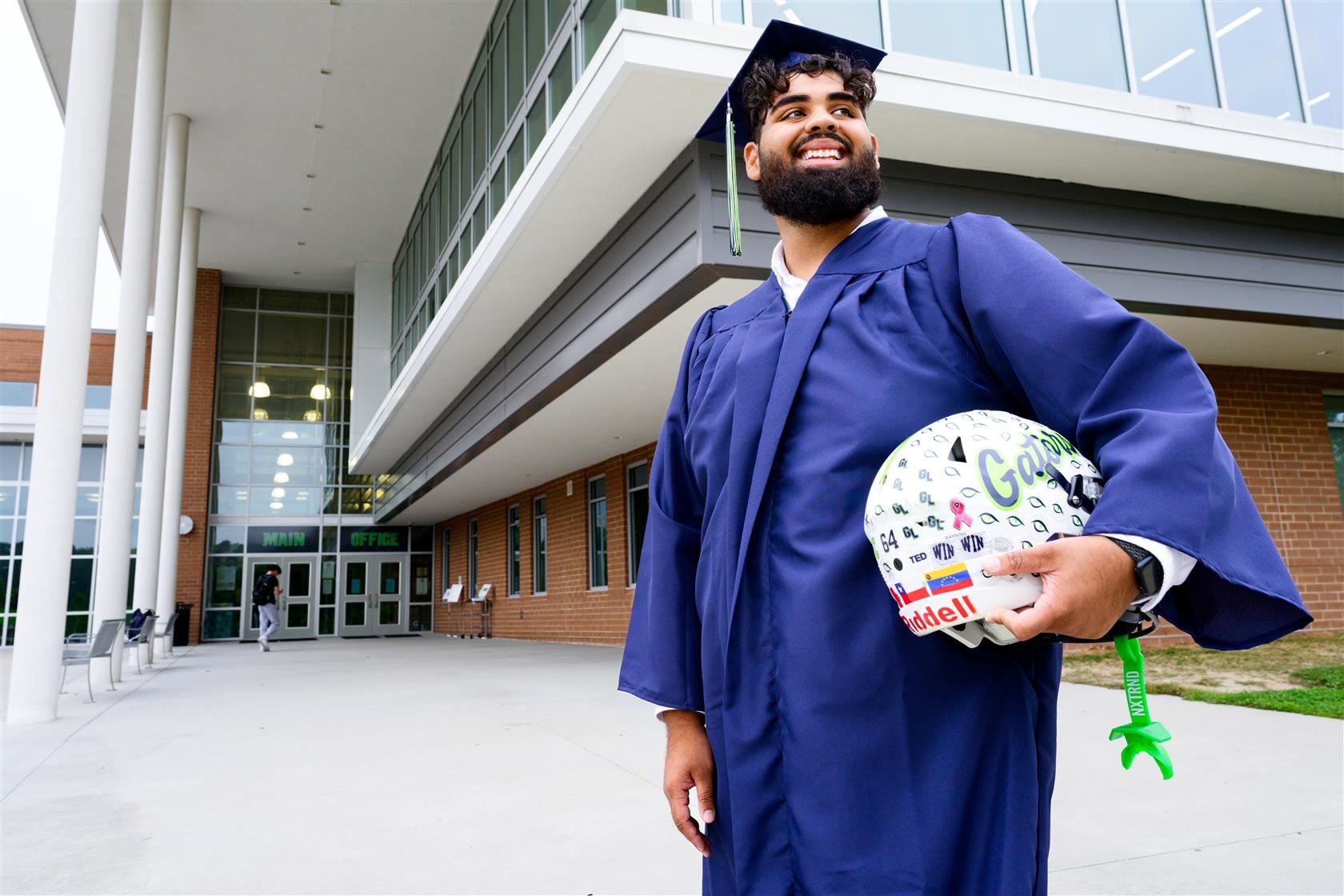 Rusho Moinuddin stands outside green level high wearing cap and gown and holding football helmet